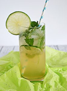 Close up of a glass of mojitos with limes and mint in the glass, a slice of lime on the rim, and a straw sticking out of it.