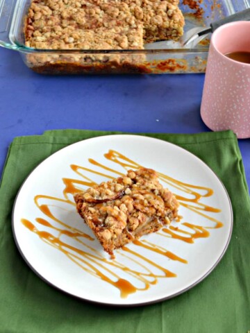 A green napkin topped by a white plate with a golden pear bar with crumble topping sitting on it with a caramel drizzle on top, a pink coffee mug to the right of it, and a pan of the bar cookies behind the plate.