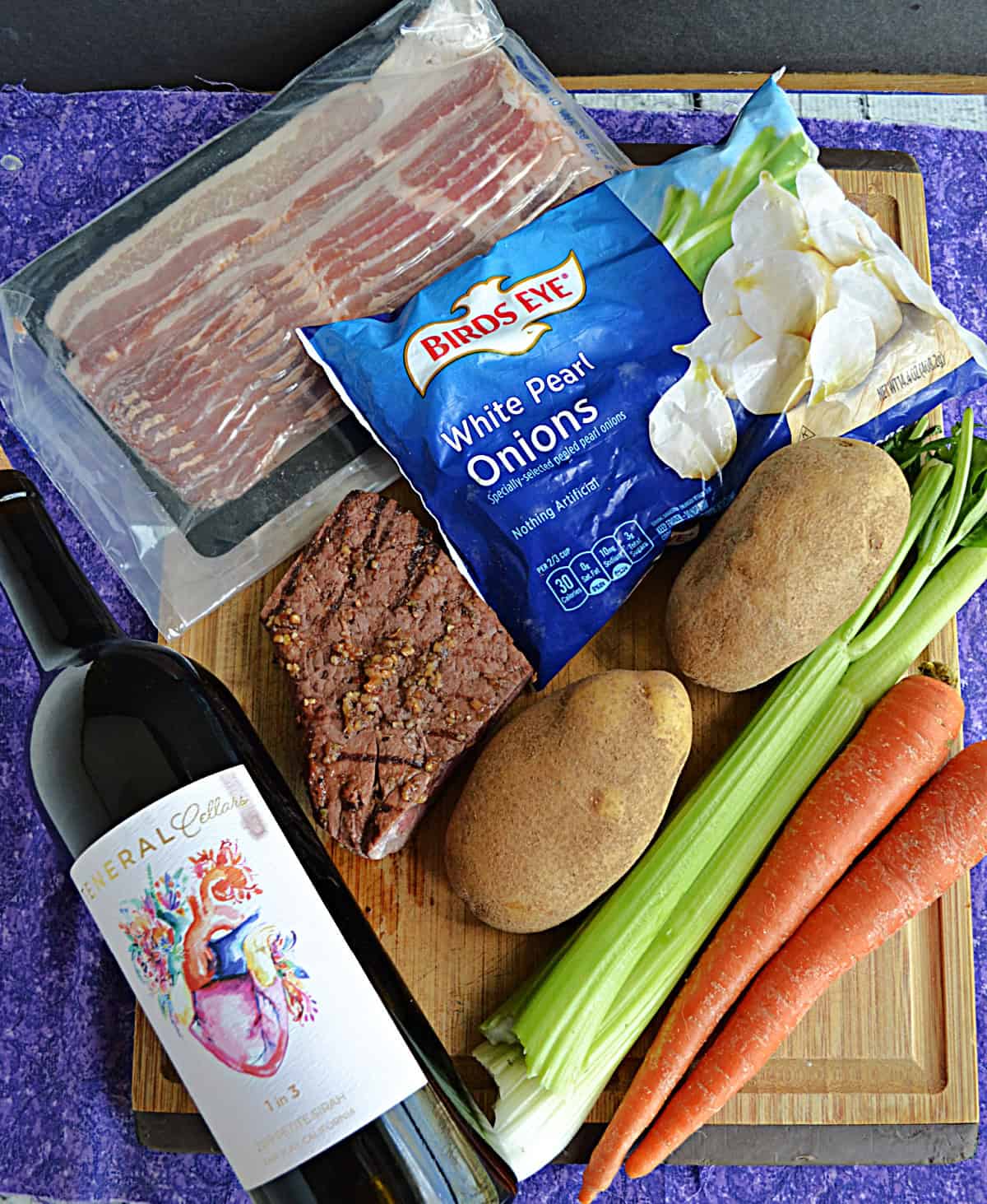 Wooden cutting board with steak, potatoes, celery, carrots, wine, and bacon on it.