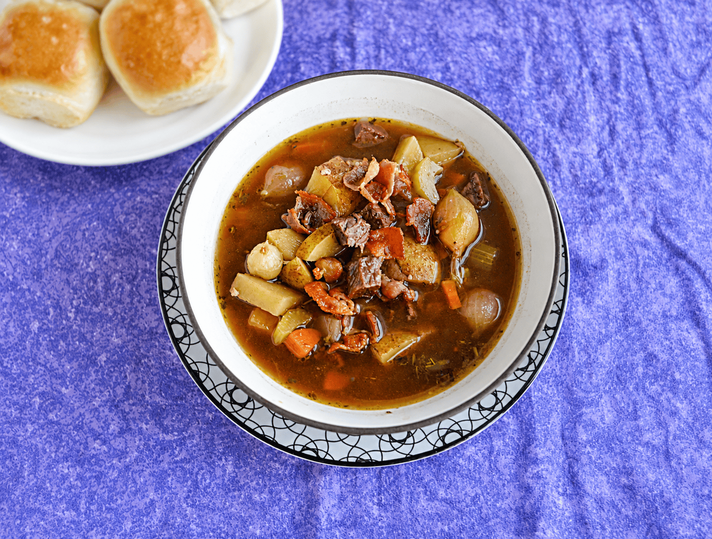 A bowl of Beef Burgundy Soup with a plate of rolls in the background.