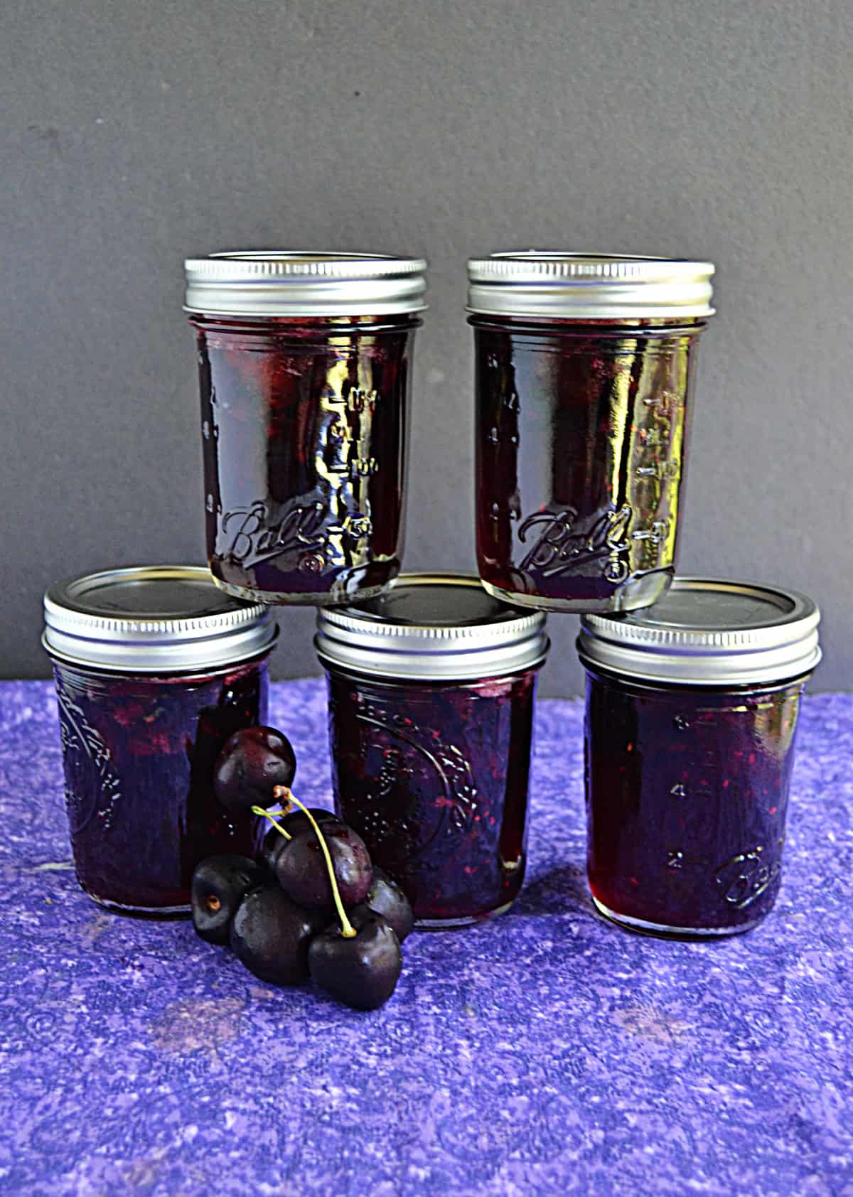 Five jars of cherry jam stacked up with a handful of cherries in from of the jars.