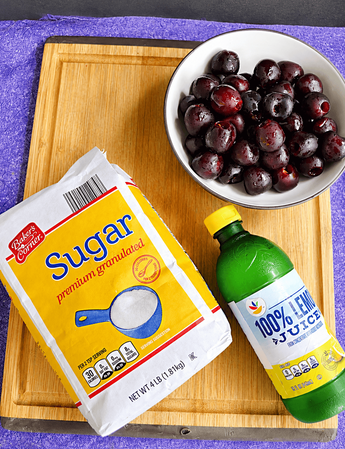 A cutting board with a bag of sugar, a bottle of lemon juice, and a bowl of cherries on it.