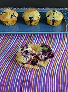 A platter with 3 blueberry muffins on it and a muffin in front with the wrapper off..