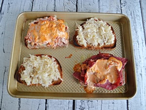 A sheet pan with four slices of bread, two with sauerkraut and cheese on them, and the other two with meat and dressing on them.
