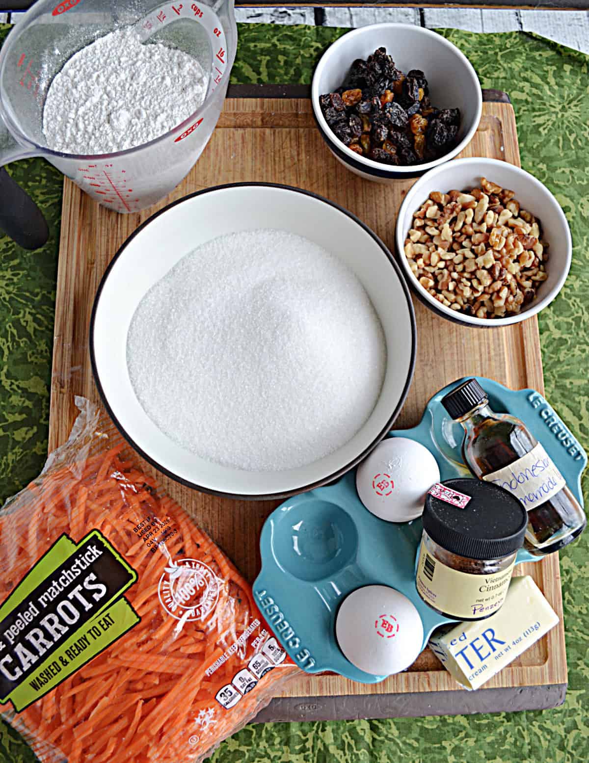 A cutting board with a bowl of sugar, nuts, raisins, carrots, and flour, 2 eggs, and spices. 