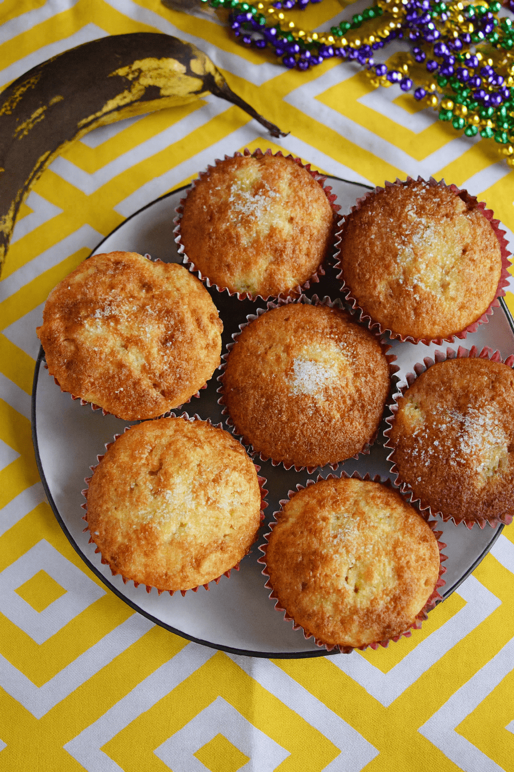 A plate of muffins with a banana behind the plate and colorful Mardi Gras beads.