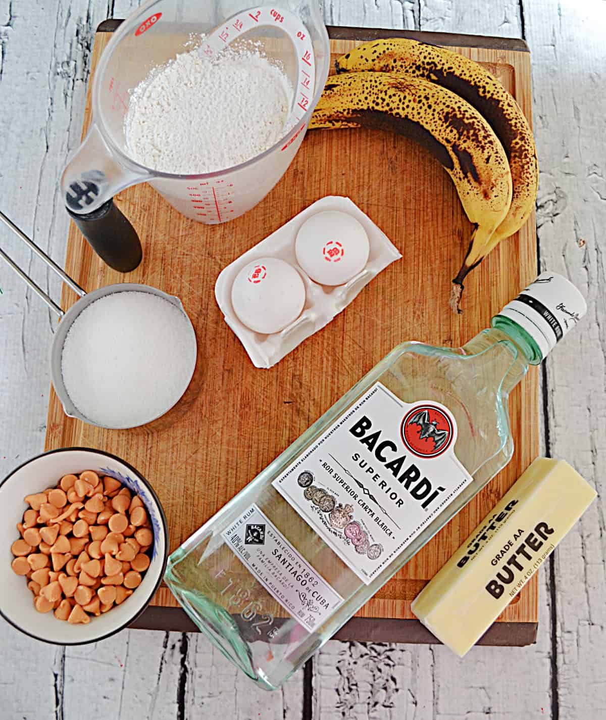 A cutting board with a banana, flour, brown sugar, butter, rum, and butterscotch chips.