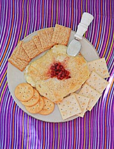 A top view of Puff pastry filled with Brie cheese and pomegranate on top with a knife stuck in it and crackers around the Brie.