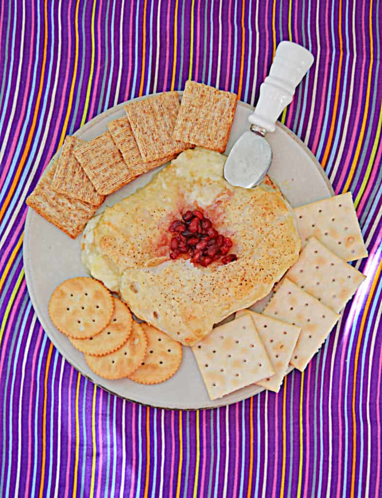 A top view of Puff pastry filled with Brie cheese and pomegranate on top with a knife stuck in it and crackers around the Brie.