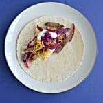 Blue background with a plate topped with a tortilla layered with beef, pickled cabbage, and sour cream.