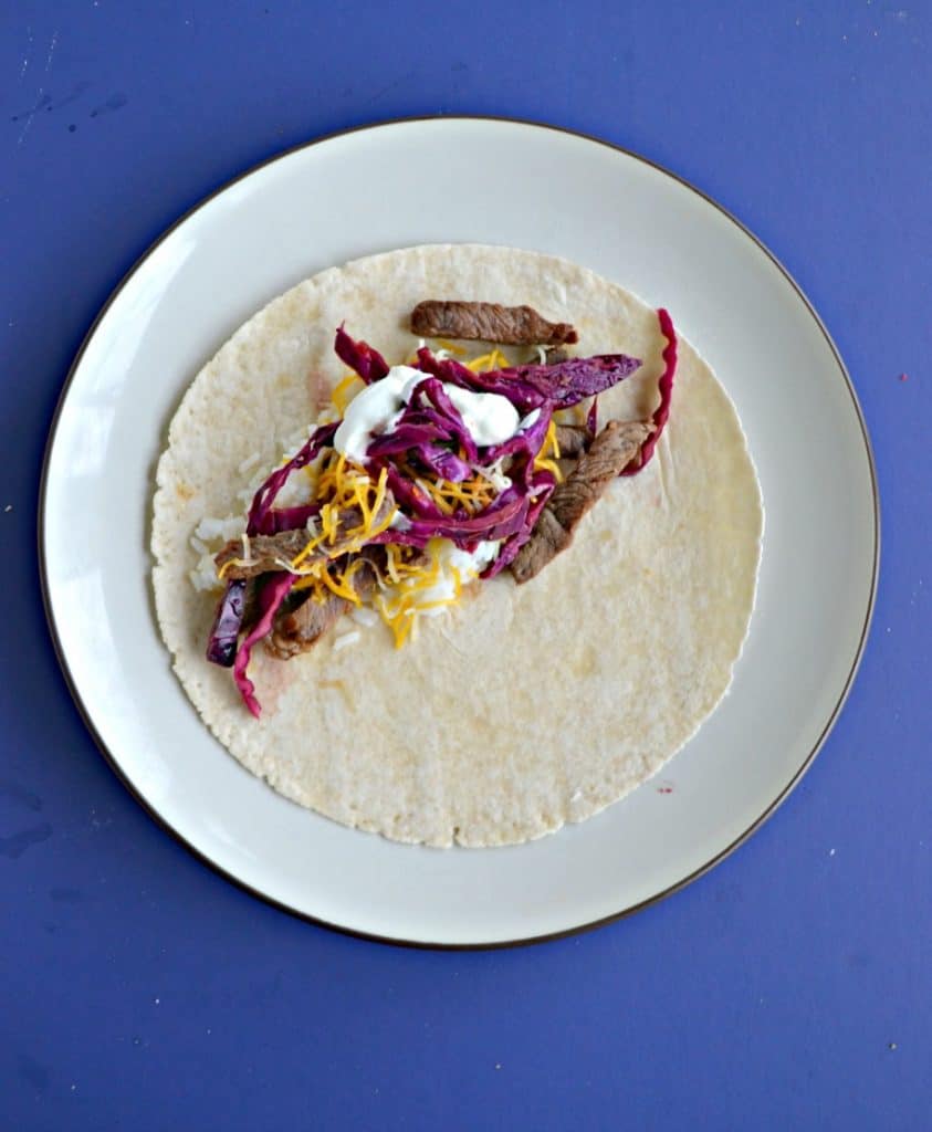 Blue background with a plate topped with a tortilla layered with beef, pickled cabbage, and sour cream.