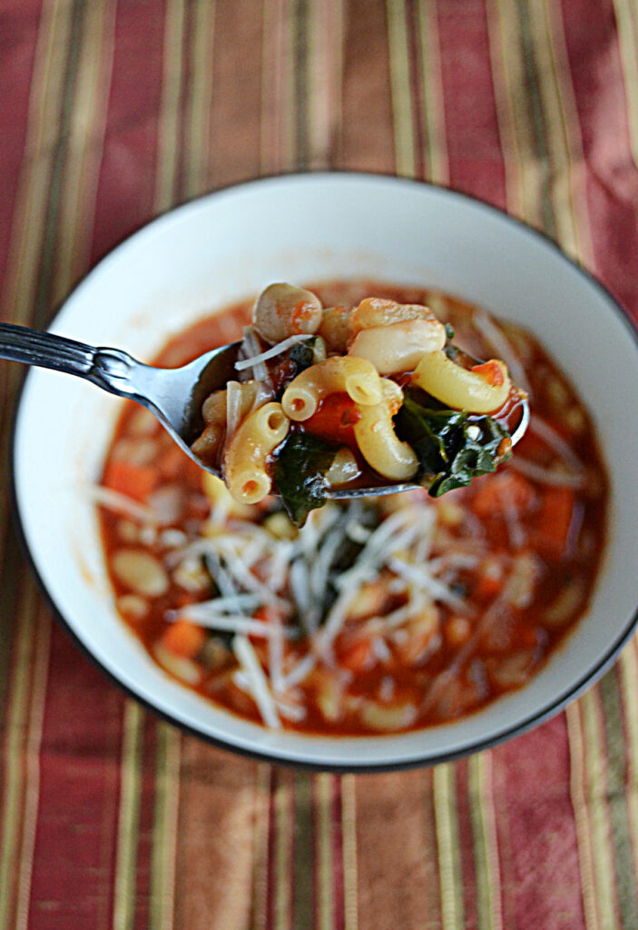A spoon with noodles, tomatoes, cheese, and spinach on it with a bowl of Pasta e Fagioli in the background. 