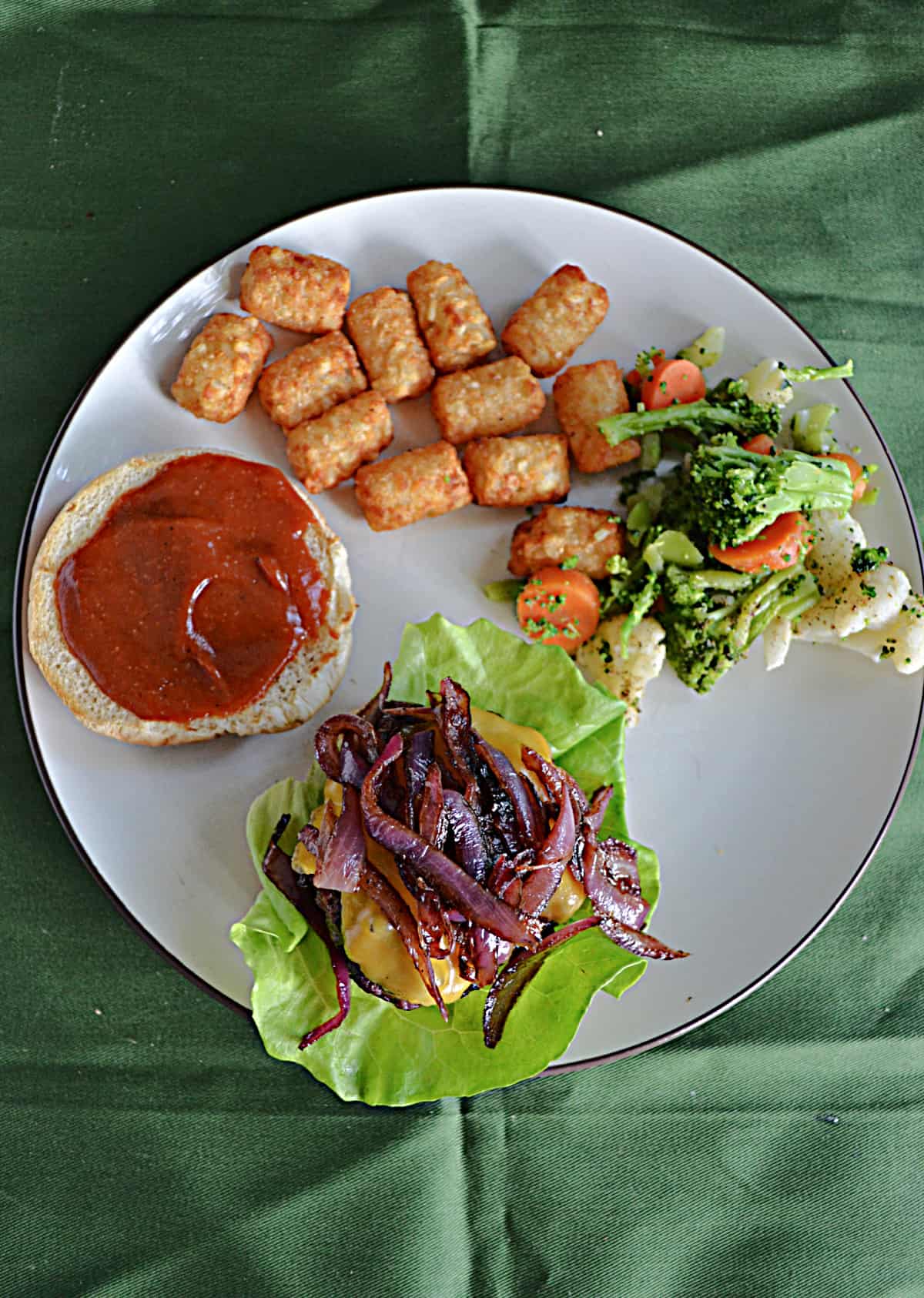 A top view of a burger with caramelized onions on top, a bun with BBQ sauce, tater tots, and mixed vegetables.  