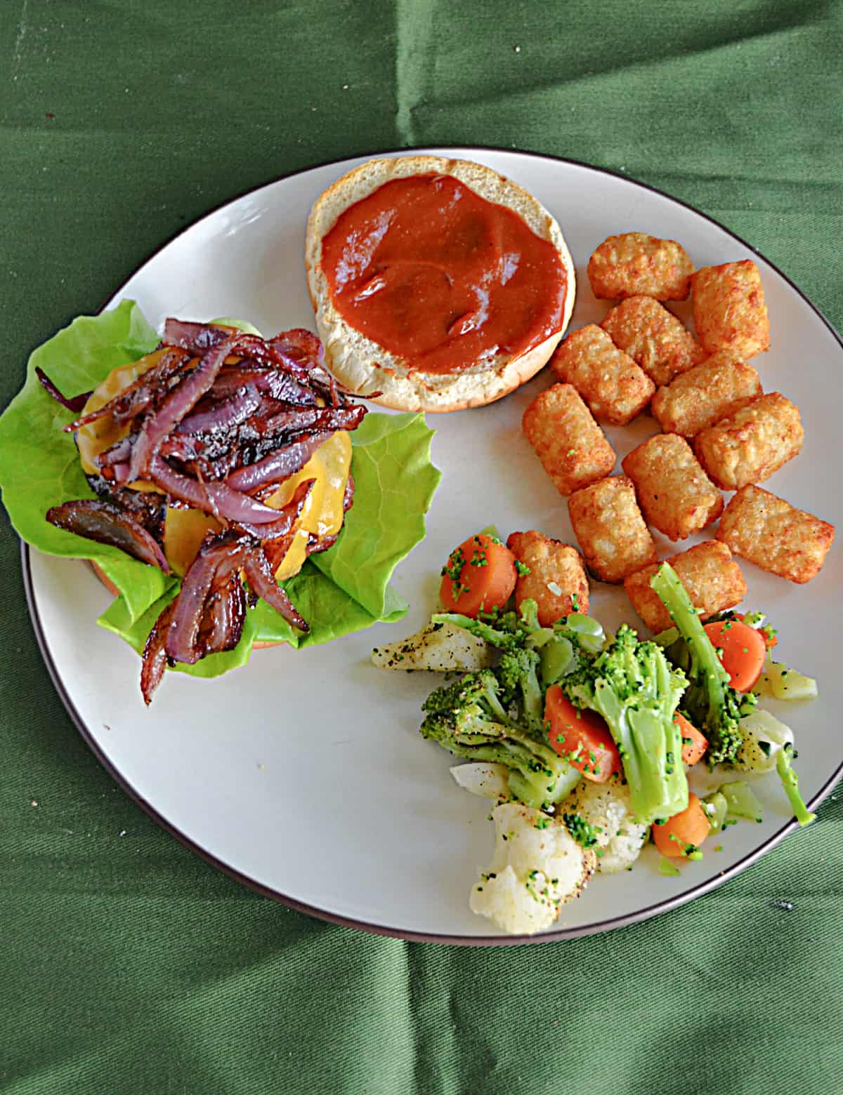A plate with a burger on a bun, a bun with bourbon BBQ sauce, tater tots, and vegetables.