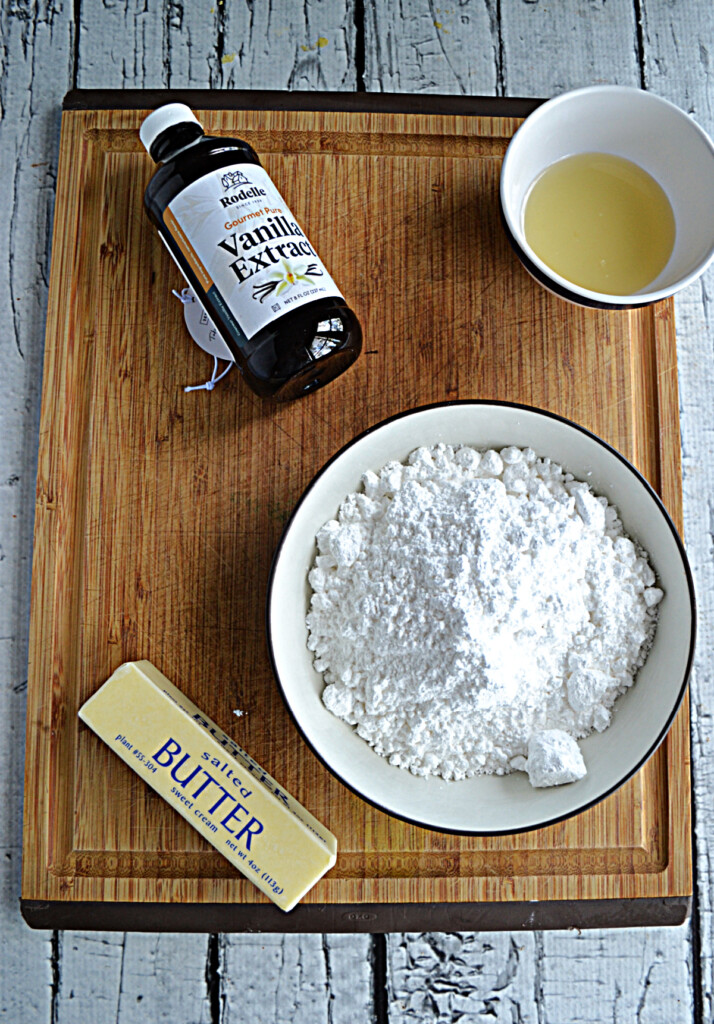 A cutting board with a stick of butter, a bowl of powdered sugar, a bowl of lemonade, and a bottle of vanilla extract.