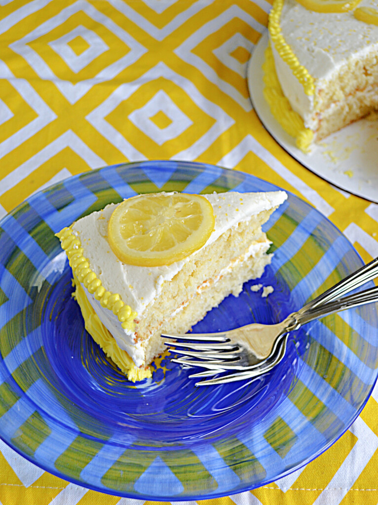 A close up of a sslice of Lemonade cake with a lemon slice on top and two forks on the plate.