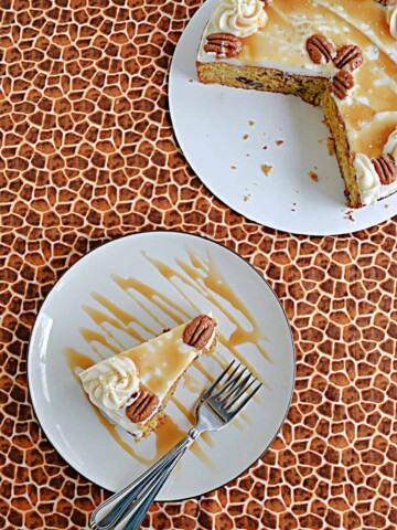 A slice of butternut squash cake topped with caramel frosting, a caramel drizzle, and pecans with two forks on the plate and the whole cake missing a slice in the corner.