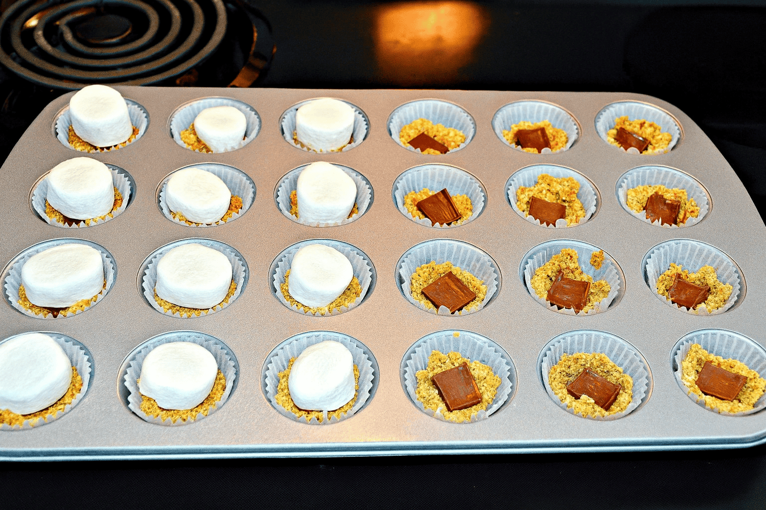 A mini muffin pan with graham cracker crust and half the cups topped with chocolate, the other half with marshmallows. 