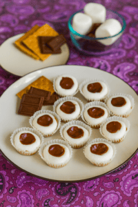 A plate of s'mores cups with graham crackers and chocolate on the plate, a bowl of marshmallows in the background.