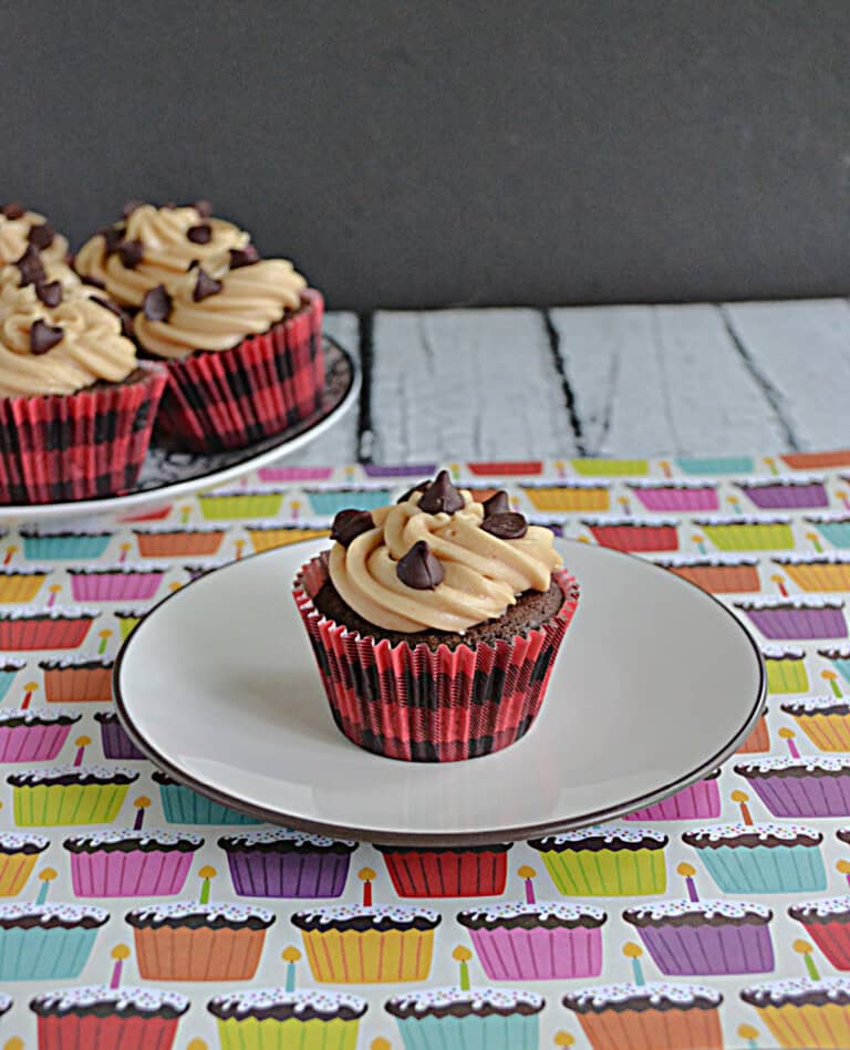 A cupcake on a plate with a tray on cupcakes in the background.