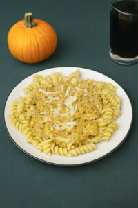 A plate of pumpkin alfredo over pasta with a glass of soda and a pumpkin in the background.