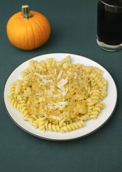 A plate of pumpkin alfredo over pasta with a glass of soda and a pumpkin in the background.