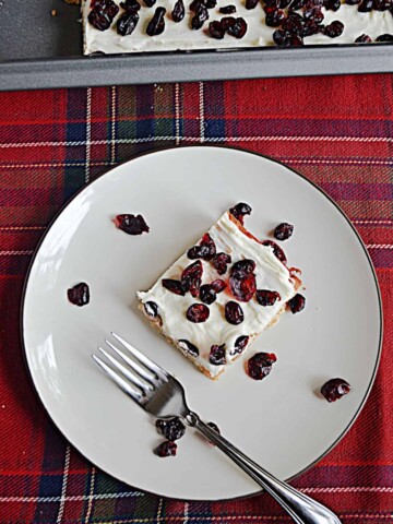 A plate with a cranberry bliss bar topped with dried cranberries, dried cranberries on the plate, a fork, and the pan of bars behind the plate.