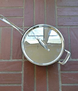 A large, silver skillet with a lid on it on top of a brick background.