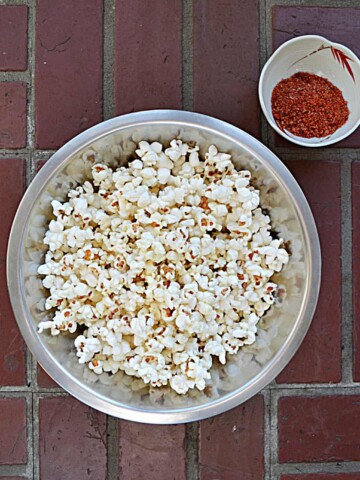 A bowl filled with fluffy white popcorn with a small bowl of red BBQ seasoning in the upper right hand corner all on a brick background.