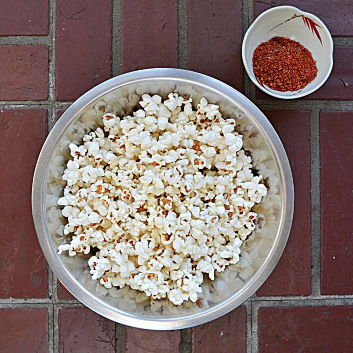 A bowl filled with fluffy white popcorn with a small bowl of red BBQ seasoning in the upper right hand corner all on a brick background.