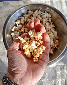 A silver bowl filled with popcorn sprinkled with red BBQ seasoning with a hand holding a handful of popcorn in the foreground on a white background.