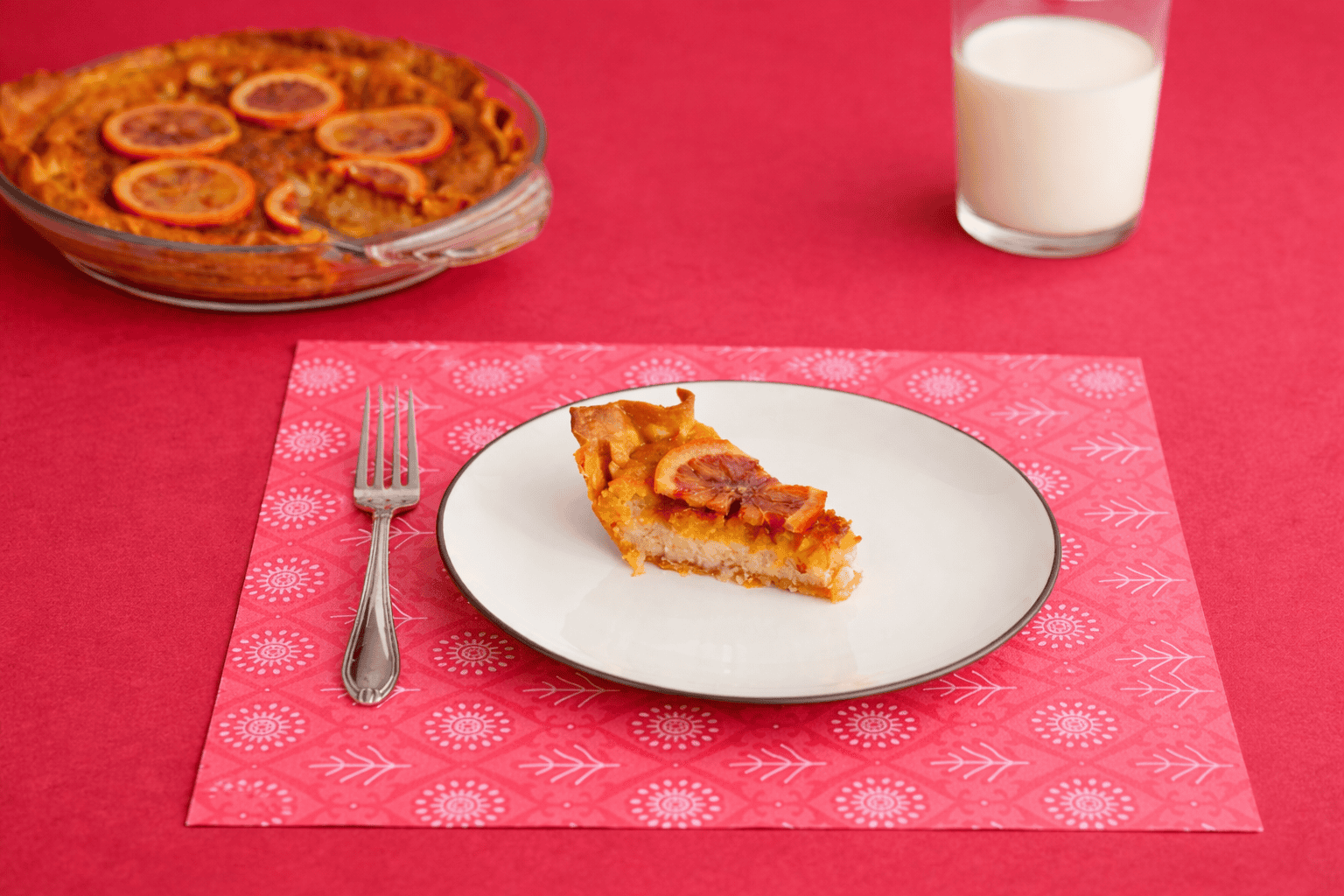 A slice of pie on a plate with the orange pie behind it along with a glass of milk.