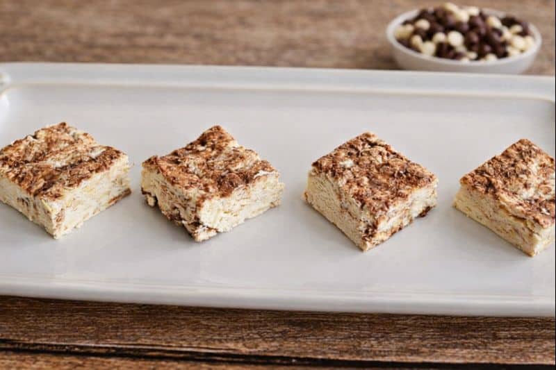 A close up of a few squares of nougat with a bowl of white and chocolate chips behind the plate.