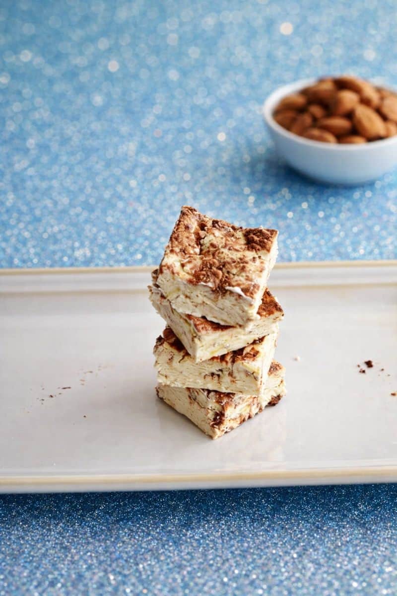 A platter with nougat squares stacked on top of each other and a bowl of almonds behind the plate.