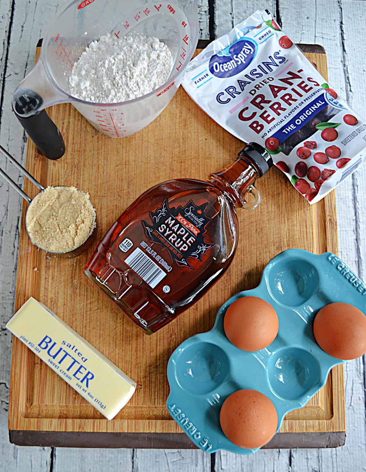 A wooden cutting board with a cup of flour, a bottle of maple syrup, 3 eggs, a stick of butter, a bag of cranberries, and a cup of brown sugr on it. 