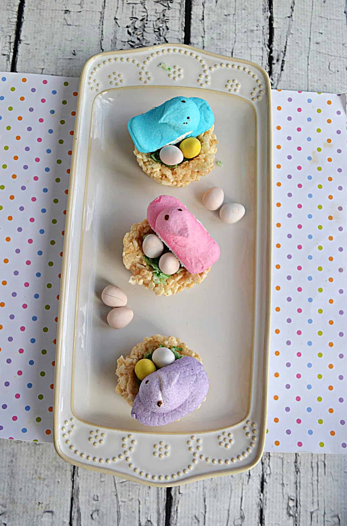 A platter with 3 bird's nests with candy eggs and Peeps on top and candy eggs around the plate.