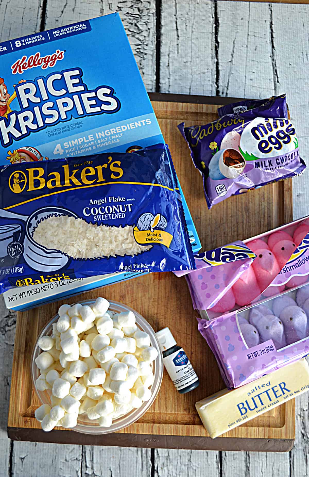 A wooden cutting board with a box of Rice Krispies, a bag of coconut, a bowl of mini marshmallows, a bags of chocolate eggs, and three boxes of Peeps.