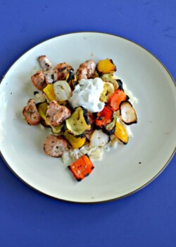 A plate piled high with white rice, grilled pork, and colorful veggies topped with a small spoonful of white Tzatziki sauce on a blue background.