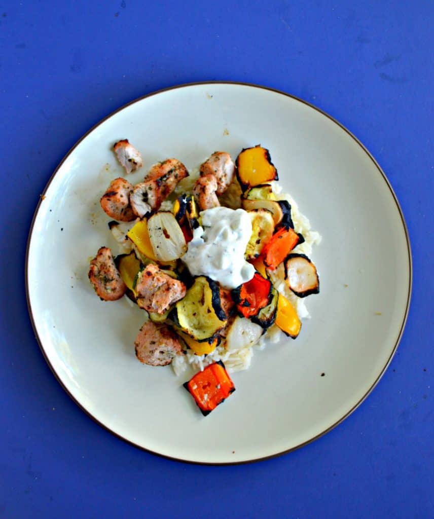 Close up image of a plate piled high with white rice, grilled pork, and colorful veggies topped with a small spoonful of white Tzatziki sauce on a blue background.