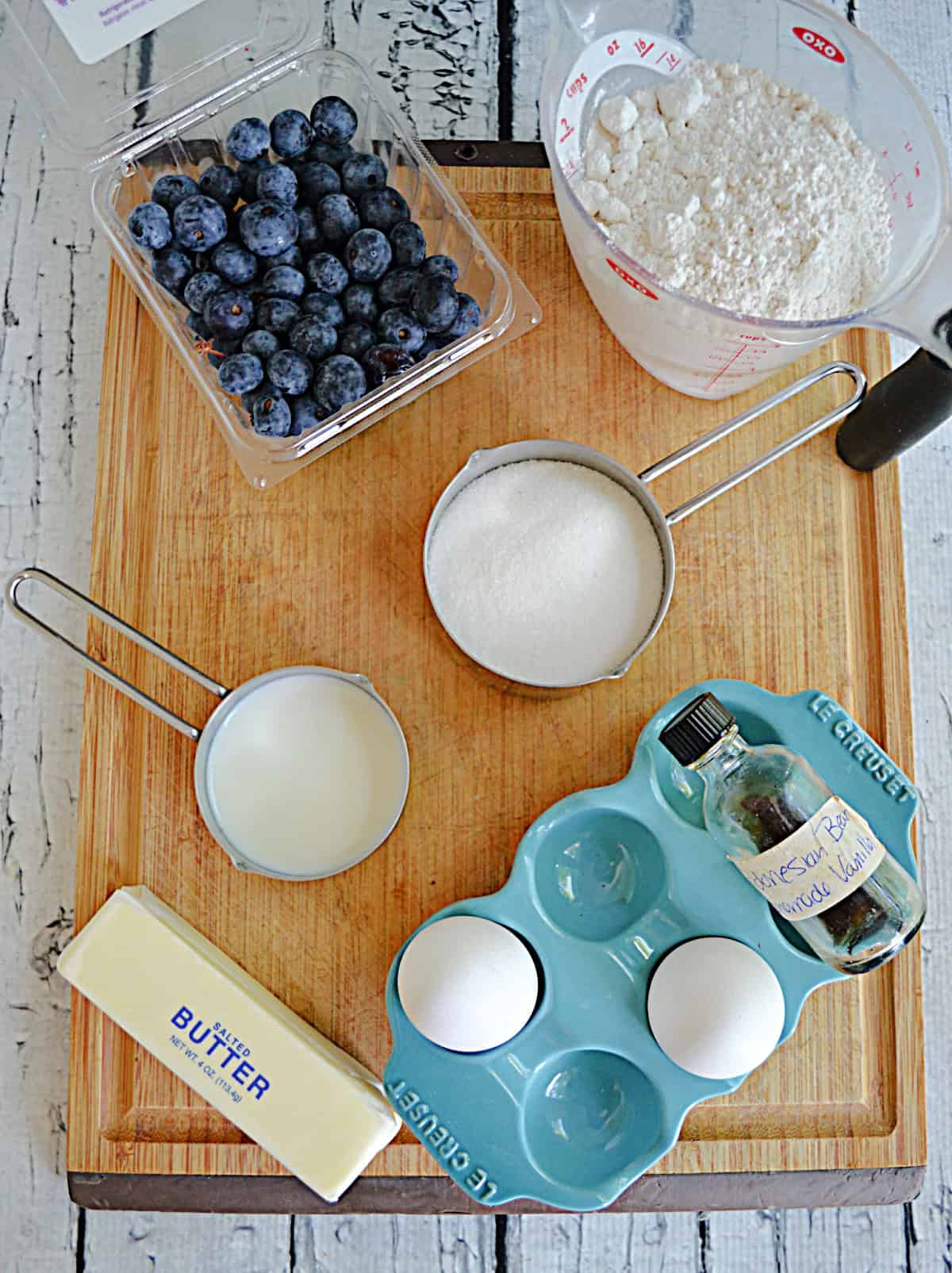 A wooden cutting board with a container of blueberries, a cup of flour, a cup of sugar, 2 eggs, and a stick of butter on it.