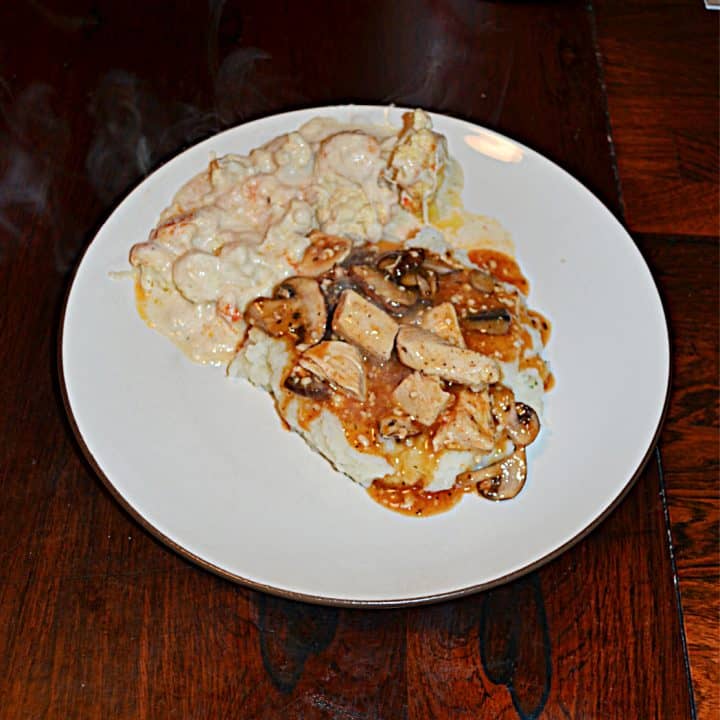 A plate with mashed potatoes topped with chicken, mushrooms, and onions in a white wine sauce and a side of cauliflower.