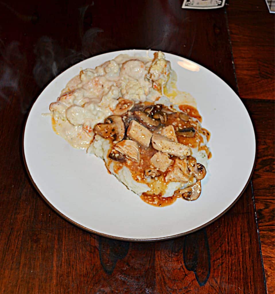 A plate with mashed potatoes topped with chicken, mushrooms, and onions in a white wine sauce and a side of cauliflower. 