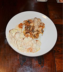 A plate with mashed potatoes topped with chicken, mushrooms, and onions in a white wine sauce and a side of creamy cauliflower.