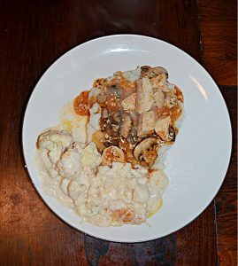 A top view of a plate with mashed potatoes topped with chicken, mushrooms, and onions in a white wine sauce and a side of cauliflower.