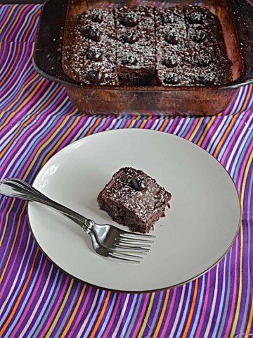 A close up view of a plate with a chocolate cranberry brownie and two forks on it with the pan of brownies behind it.