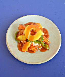 A plate piled high with green peppers, chicken, pineapple, and red sweet and sour sauce with a pineapple ring on top on a blue background.