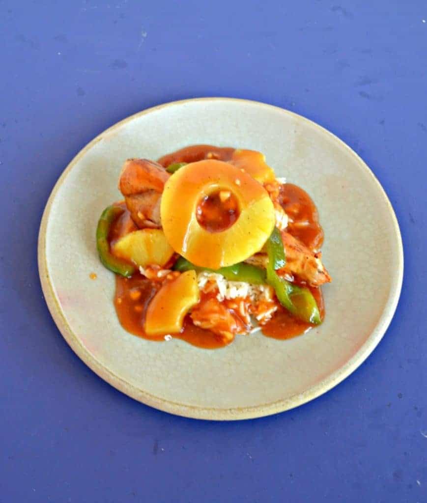 A plate piled high with green peppers, chicken, pineapple, and red sweet and sour sauce with a pineapple ring on top on a blue background.