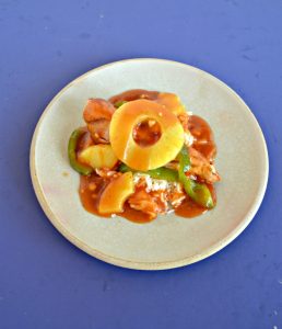 Angled view of a plate piled high with green peppers, chicken, pineapple, and red sweet and sour sauce with a pineapple ring on top on a blue background.