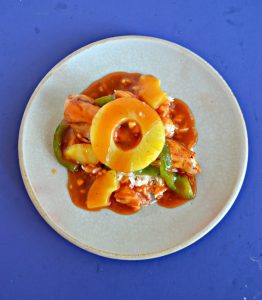 Top view of a plate piled high with green peppers, chicken, pineapple, and red sweet and sour sauce with a pineapple ring on top on a blue background.