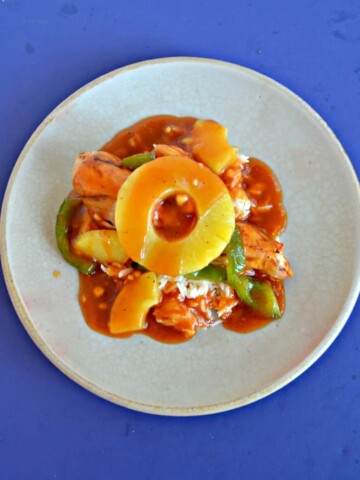 Top view of a plate piled high with green peppers, chicken, pineapple, and red sweet and sour sauce with a pineapple ring on top on a blue background.